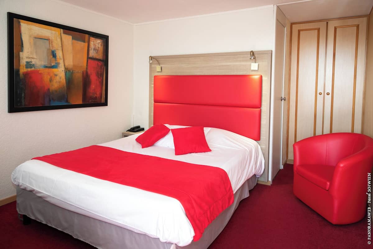 A hotel room with a large bed, a red chair, a painting on the wall, and a wooden wardrobe.
