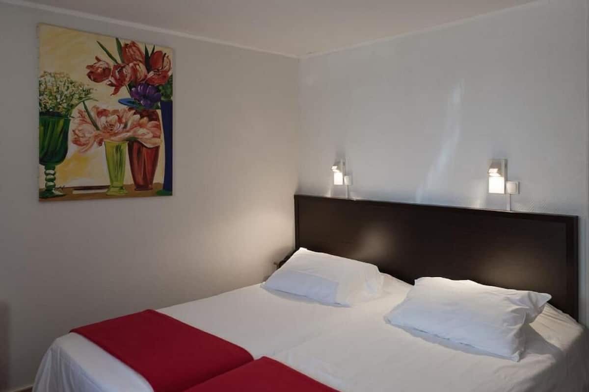A hotel room with a bed, two pillows, a red and white bedspread, and a painting of flowers on the wall.