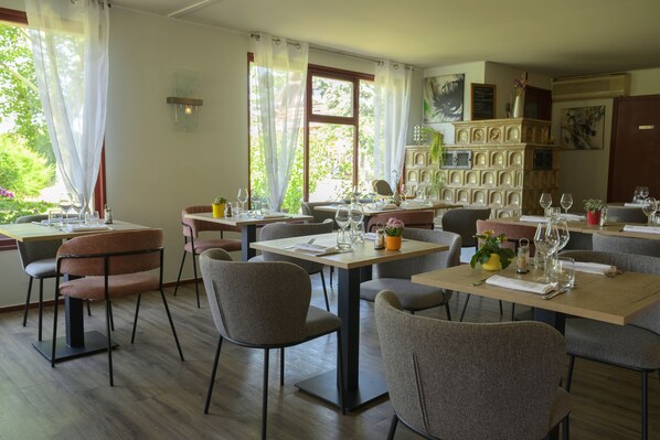 A dining area with multiple tables and chairs, a fireplace with decorative tiles, and large windows with curtains.