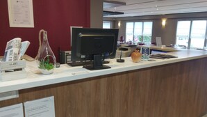 Een receptiebalie met een computer, een telefoon en een kleine plant. Er hangen borden aan de muur en er is een raam op de achtergrond.