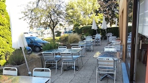 Een terras buiten met metalen tafels en stoelen, omringd door groen en geparkeerde auto's.