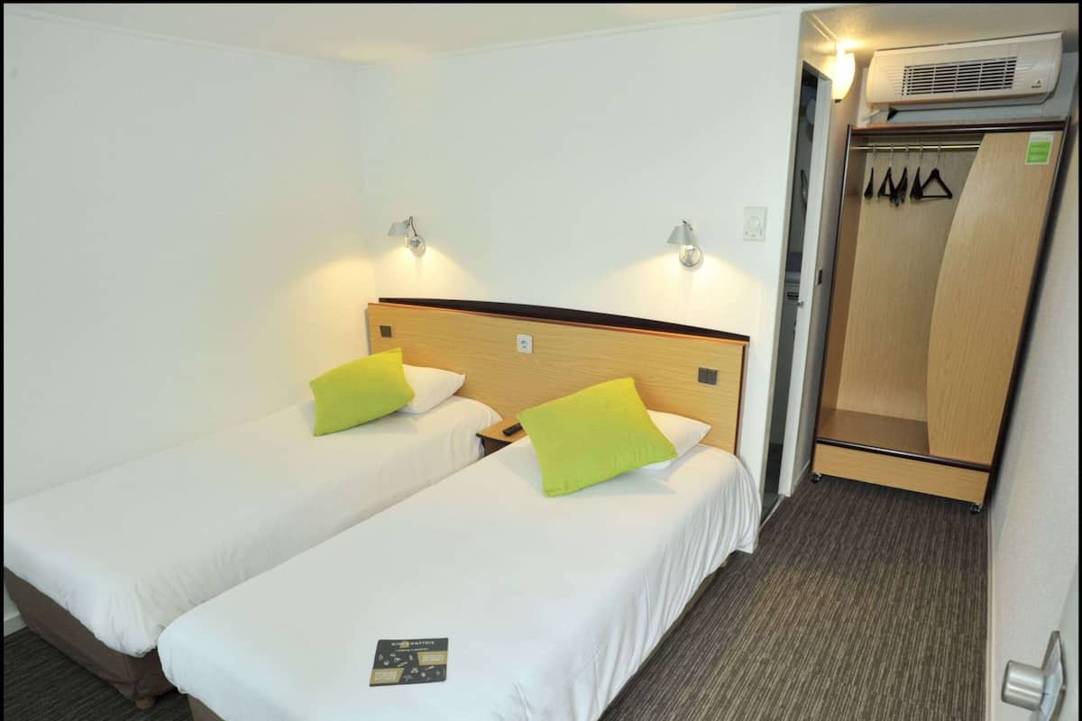 Une chambre d’hôtel avec deux lits simples, une tête de lit en bois, une petite table de chevet, une commode, une lampe fixée au mur et une armoire avec un miroir.