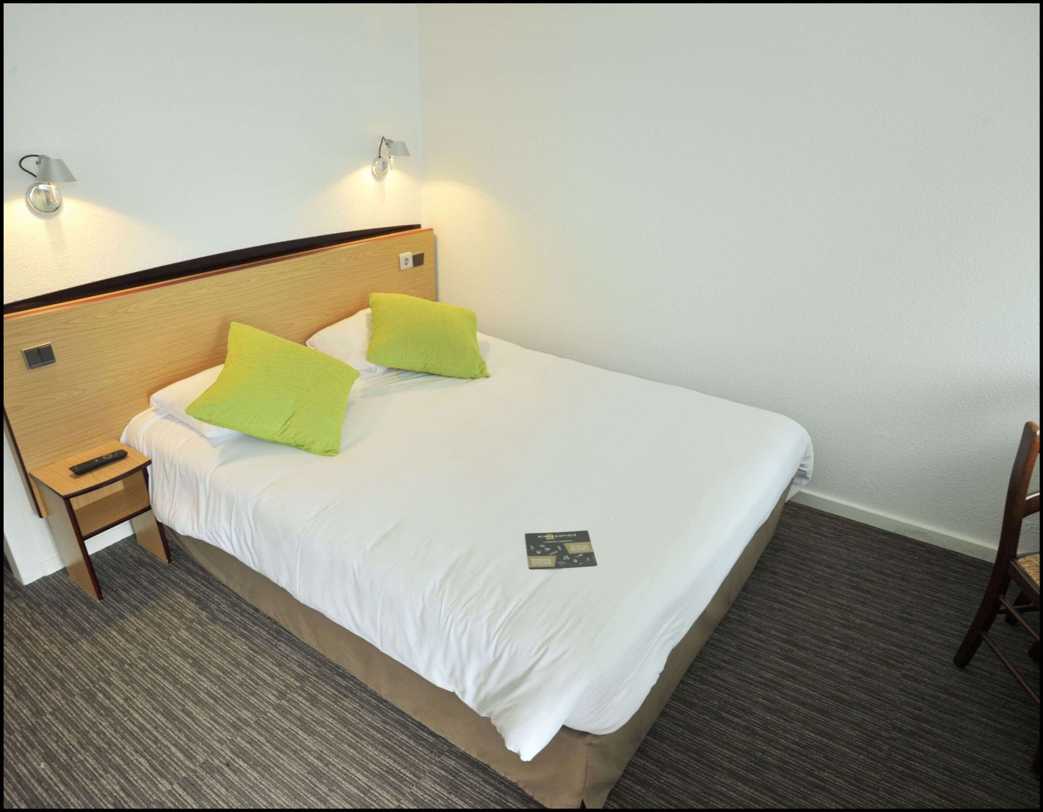 A single bed with a wooden headboard, two green pillows, a white bedsheet, a remote control on a wooden nightstand, and a small card on the bed.