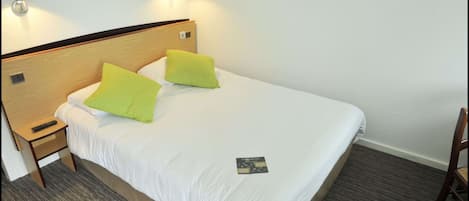 A single bed with a wooden headboard, two green pillows, a white bedsheet, a remote control on a wooden nightstand, and a small card on the bed.