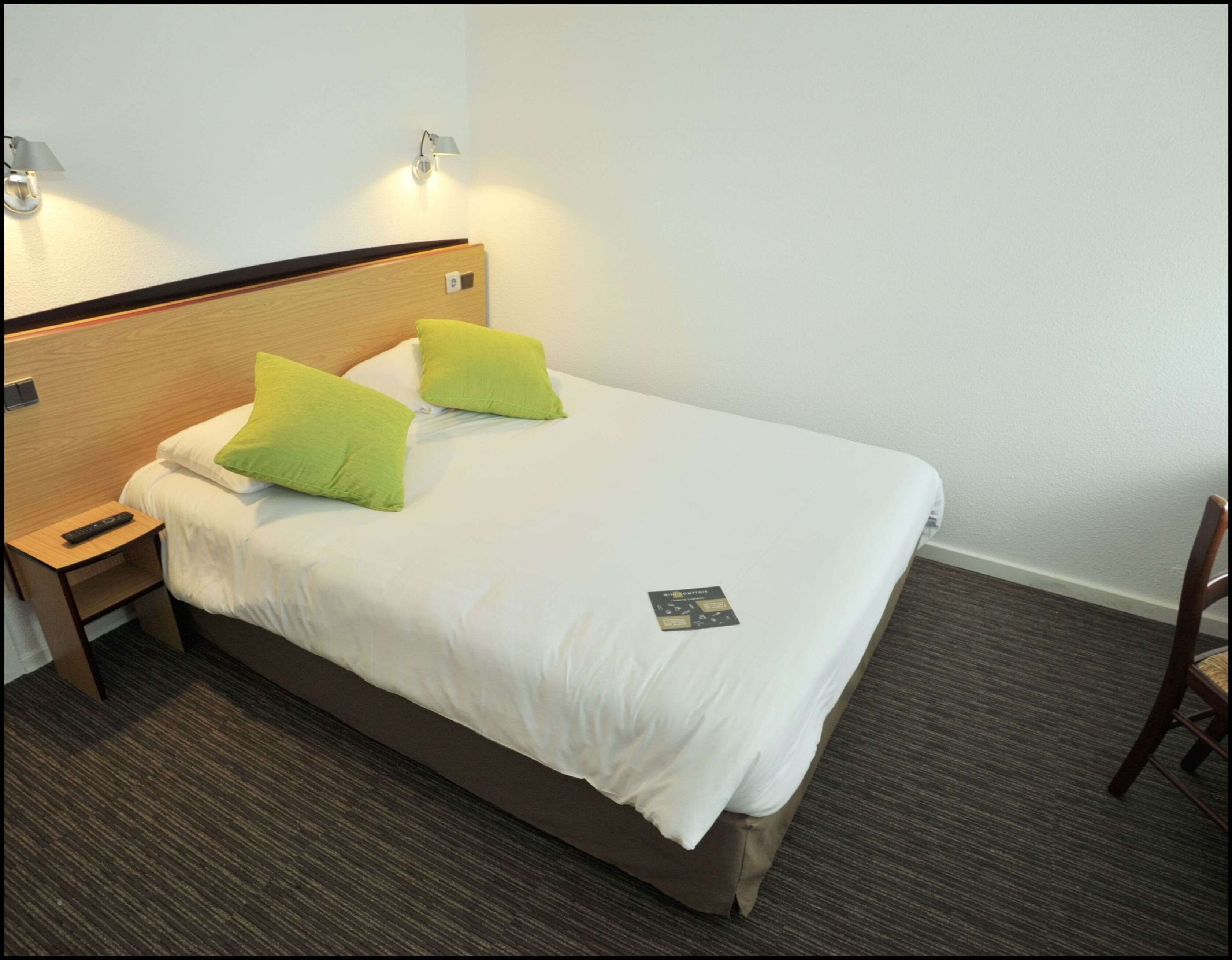 Una cama bien hecha con ropa de cama blanca y dos almohadas verdes, una mesita de noche con un control remoto y una lámpara fijada a la pared.