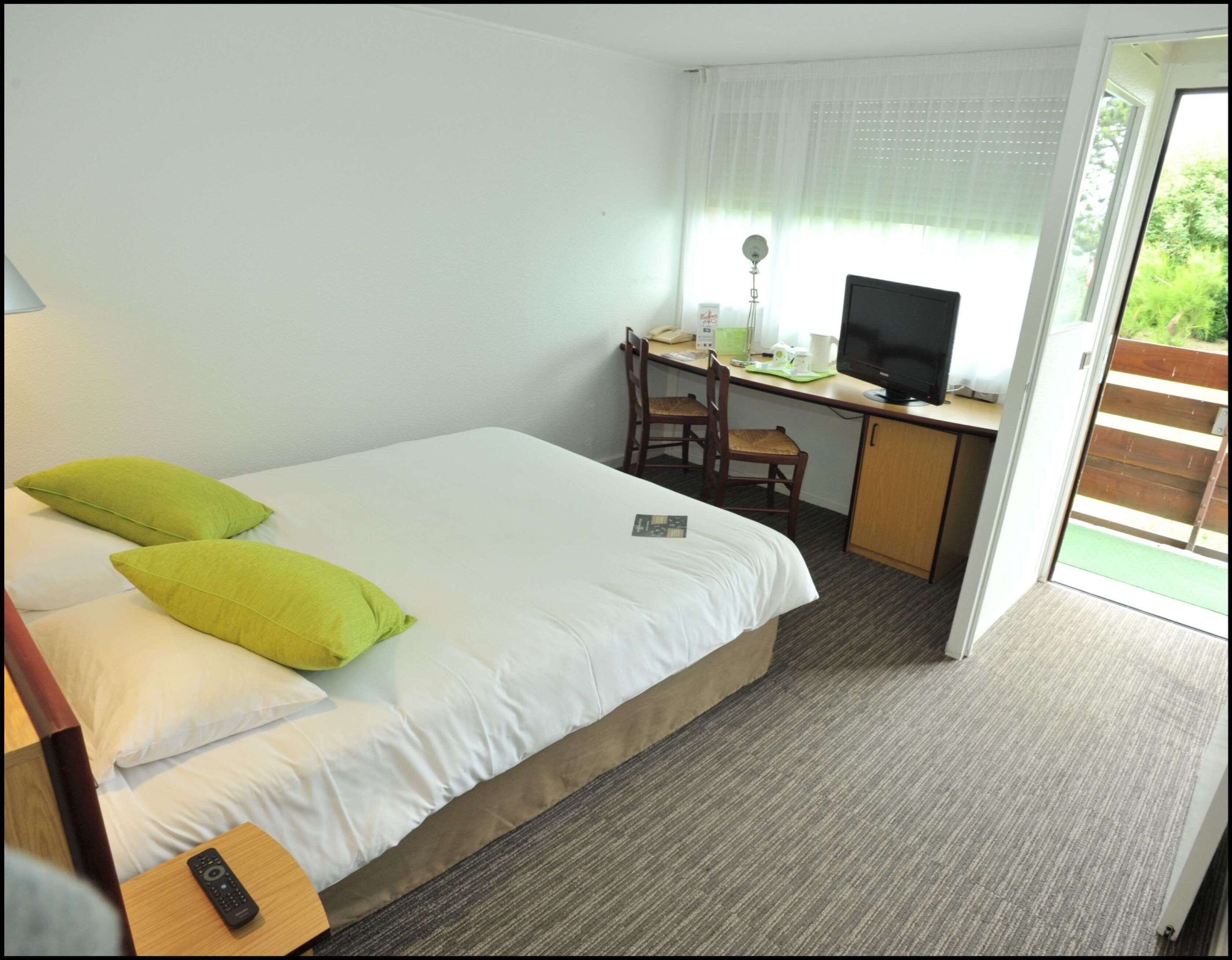 Habitación de hotel con una cama, un escritorio con computadora y televisor, y ventana con vista a follaje.