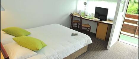 Habitación de hotel con una cama, un escritorio con computadora y televisor, y ventana con vista a follaje.