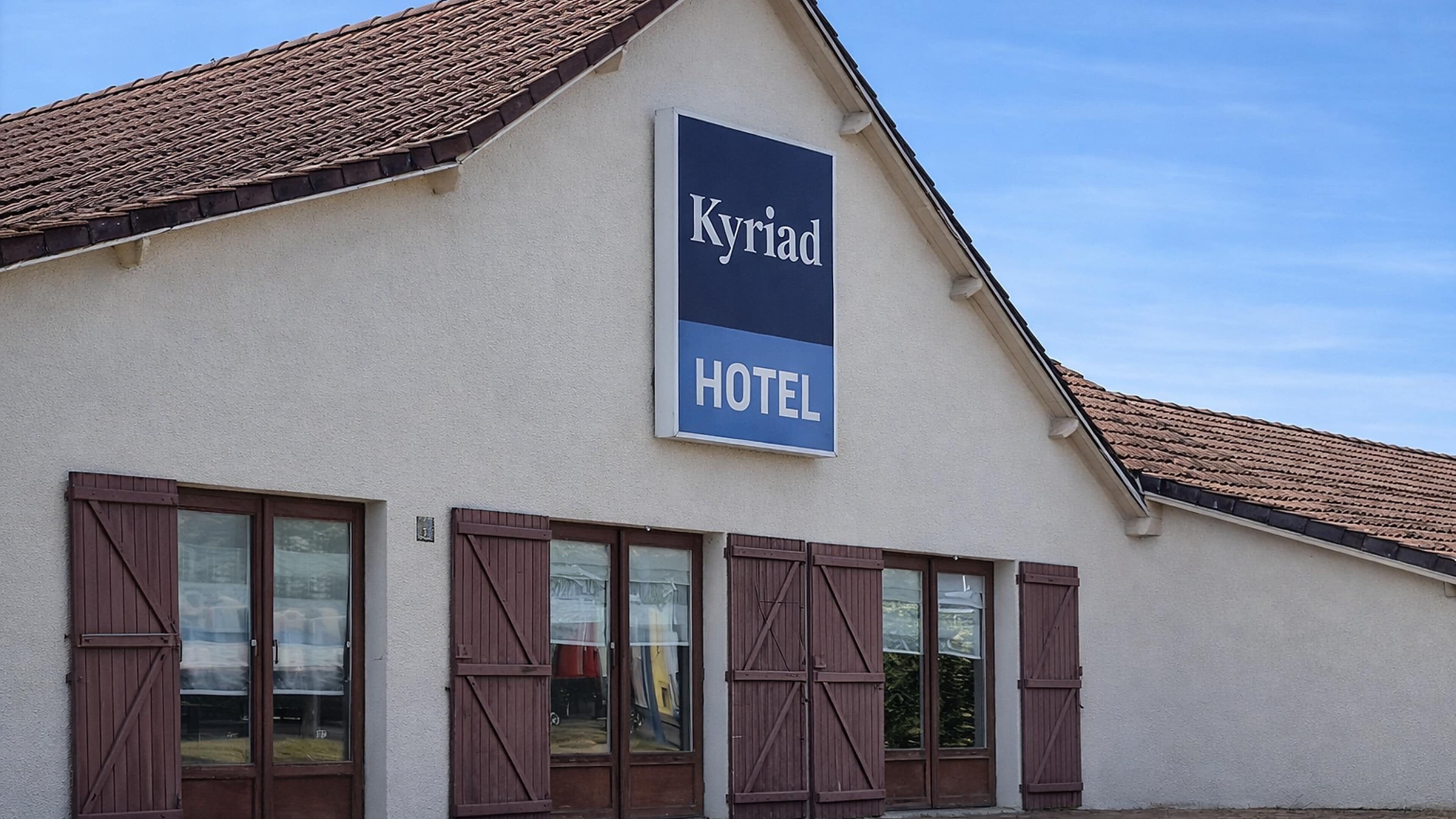 Un edificio con un cartel que dice 'Kyriad HOTEL' y amplias ventanas con contraventanas.