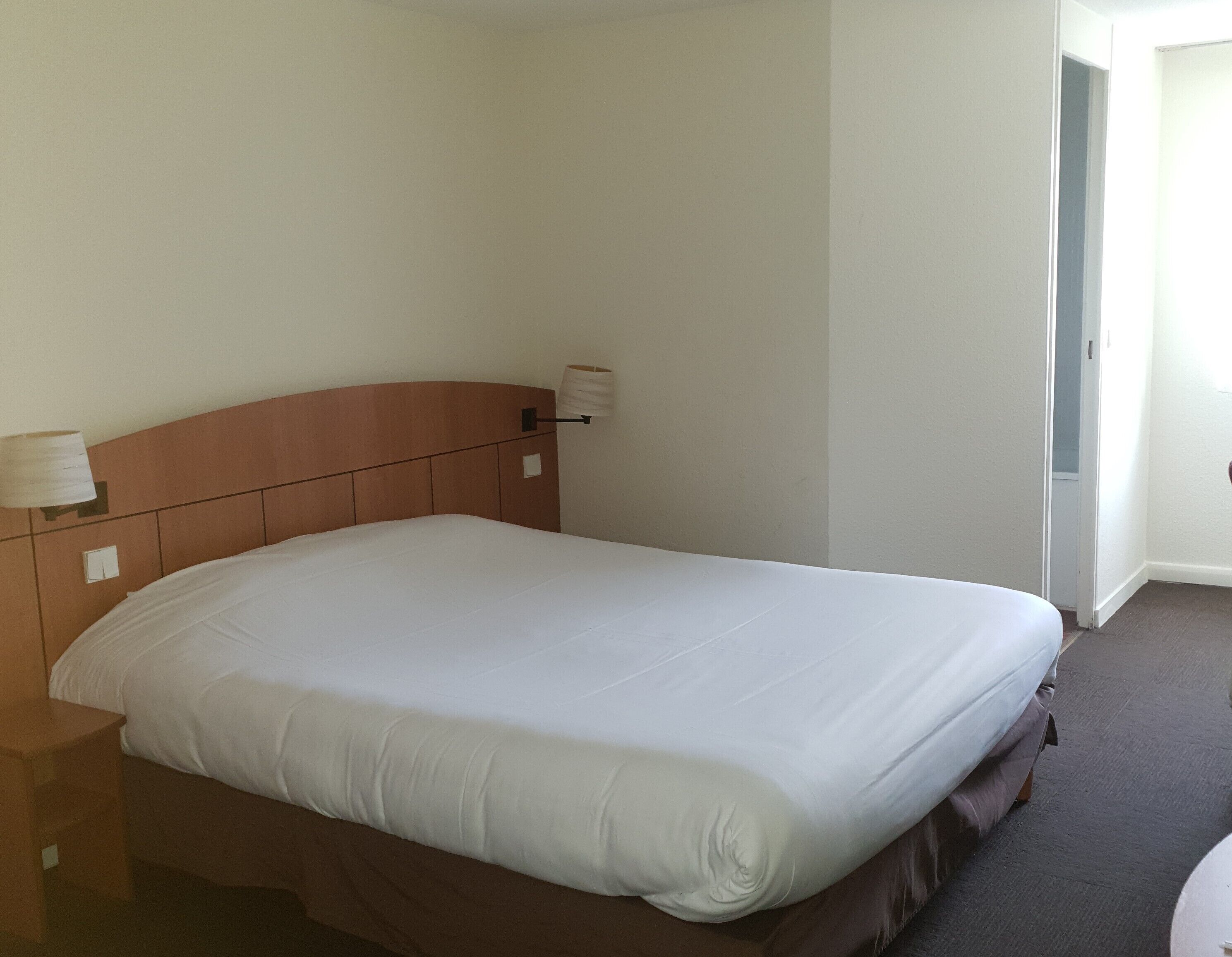 A neatly made bed in a hotel room with a wooden headboard, a bedside lamp, and a door to the right.