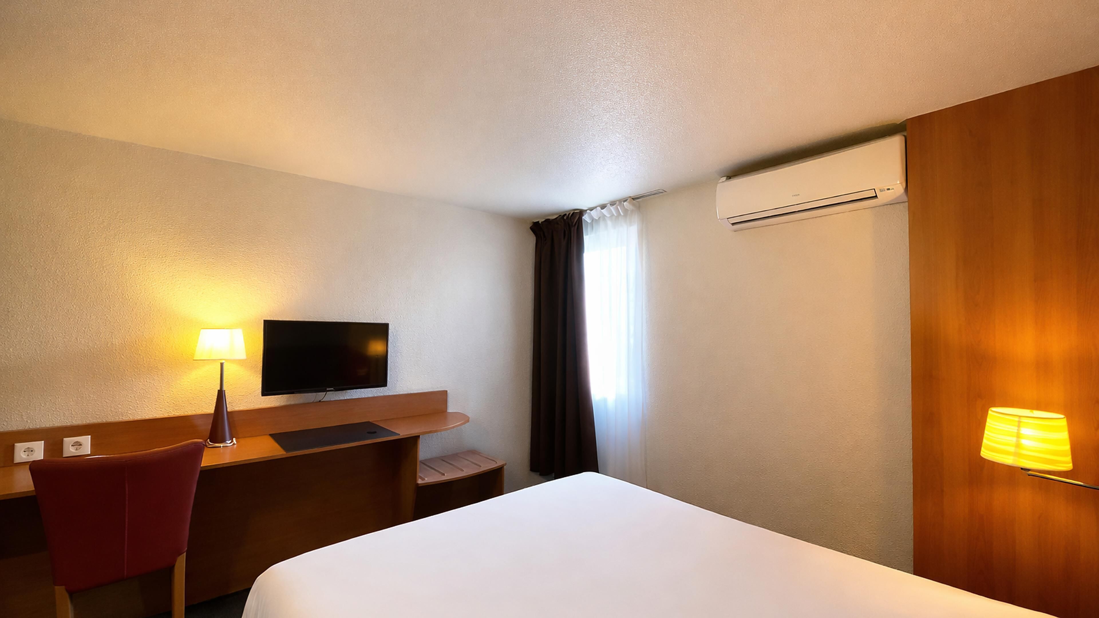 Une chambre d’hôtel équipée d’un lit, d’un bureau, d’une chaise, d’une télévision, d’une lampe, d’un climatiseur et de rideaux.