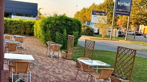 An outdoor seating area with wicker chairs and tables, a hedge, and a sign for 'Kyriad Restaurant' in the background.