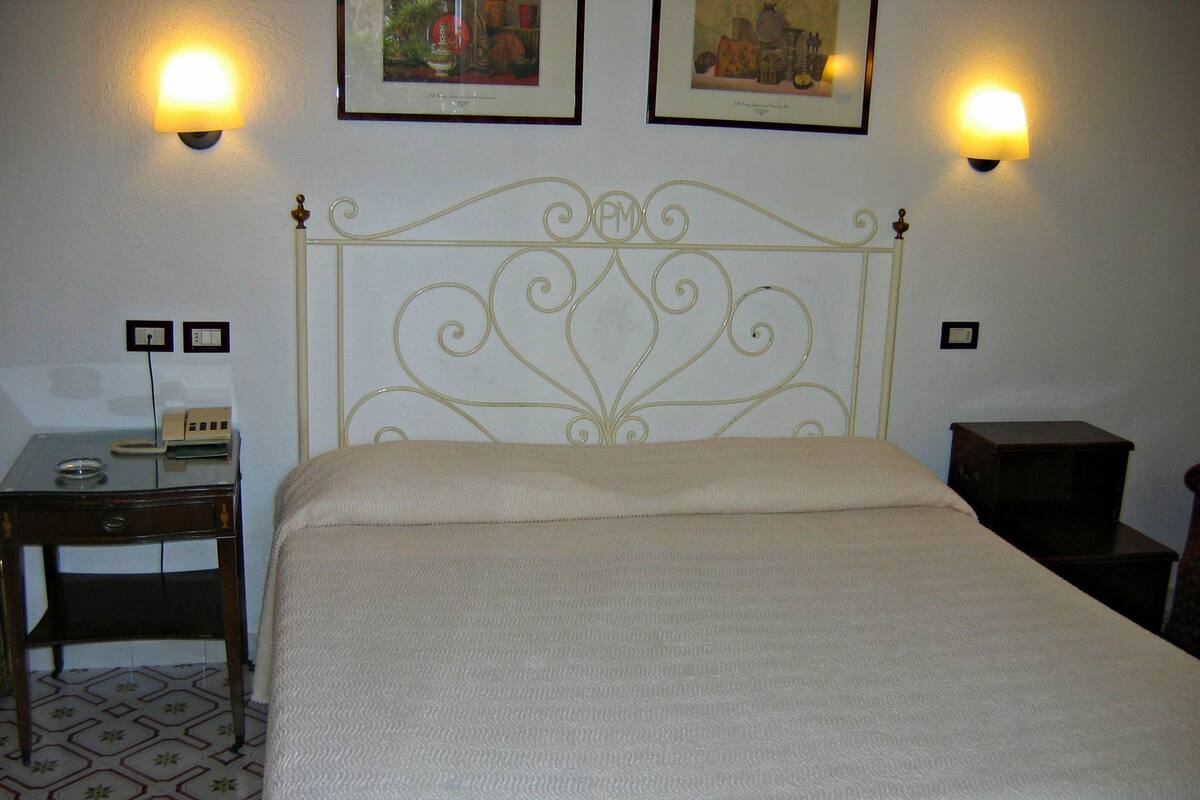 Un lit bien fait, avec une tête de lit, deux tables de chevet, un petit bureau et deux lampes fixées au mur.