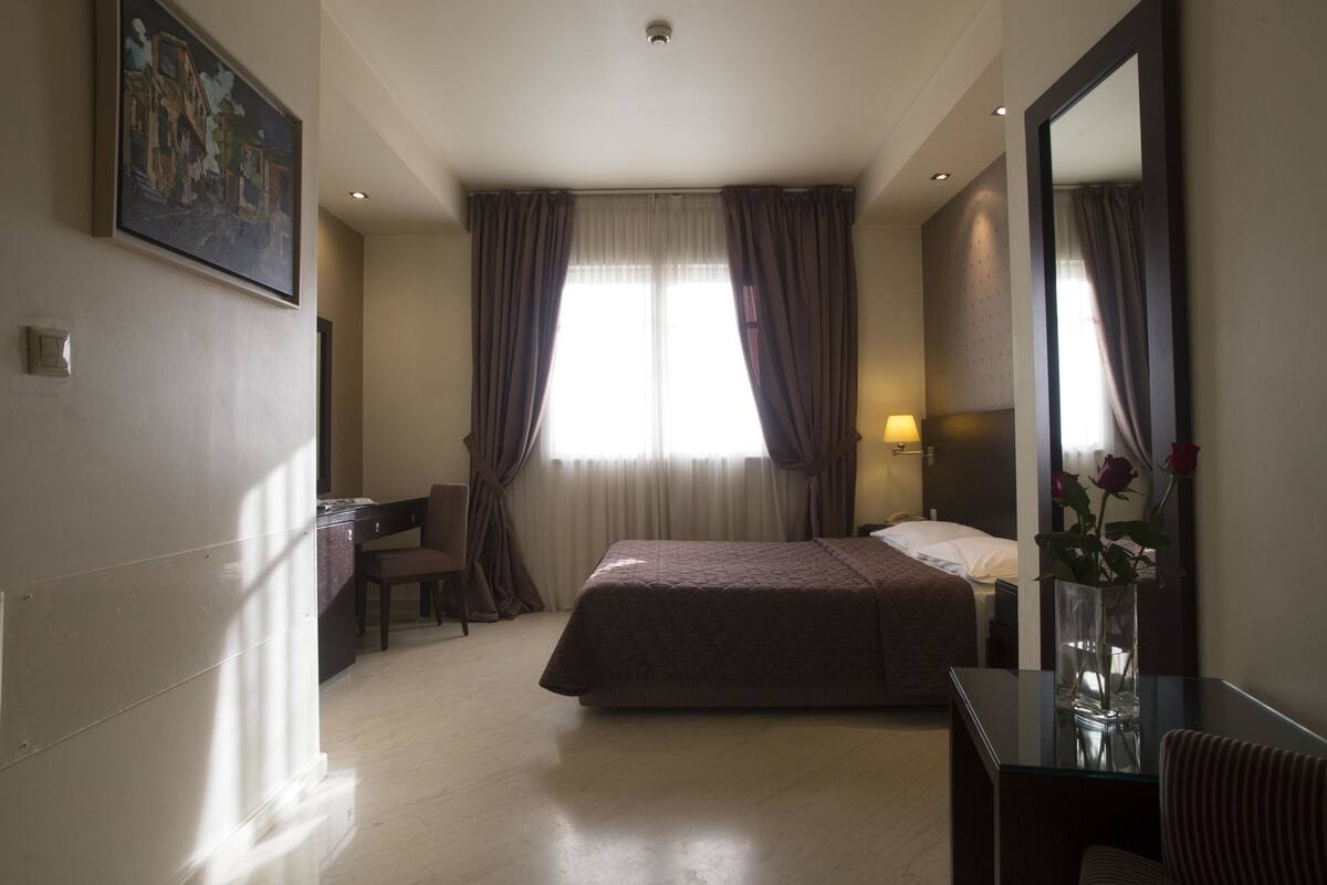 Een hotelkamer met een bed, een bureau, een stoel, een nachtkastje, een lamp, een spiegel en een schilderij aan de muur.