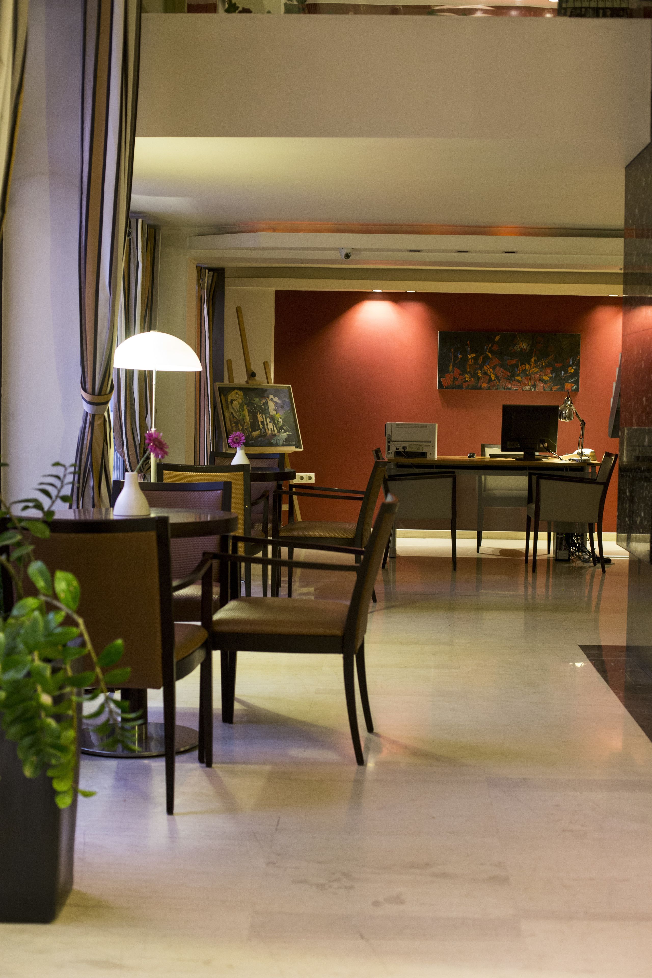 A hotel lobby with a reception desk, chairs, a table with a lamp, and a potted plant.