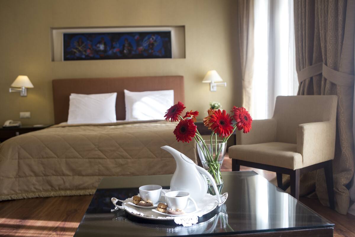 Een hotelkamer met een bed, een stoel, een dienblad met kopjes en gebak, en een vaas met bloemen.