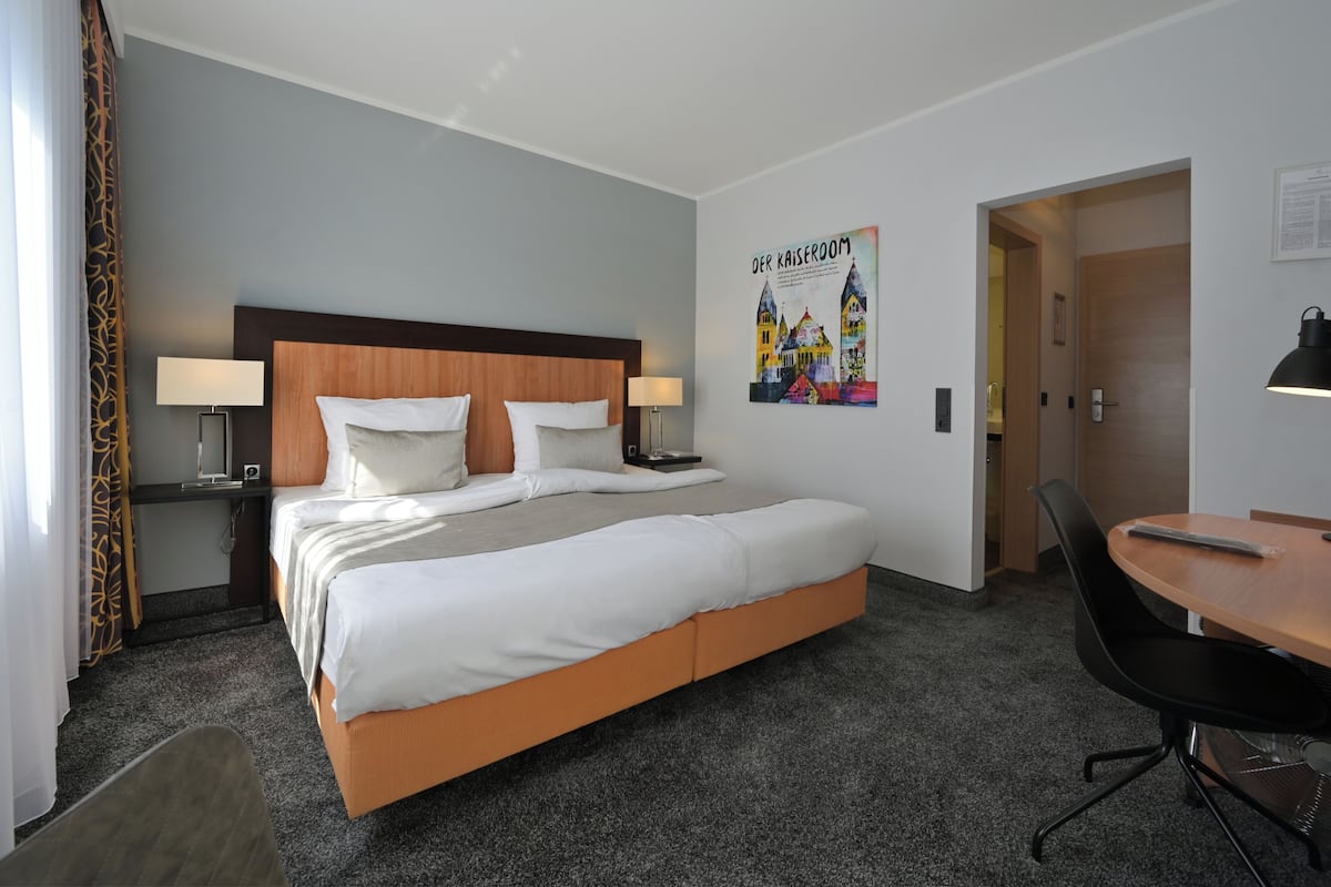 Une chambre d’hôtel avec un grand lit, deux tables de chevet avec des lampes, un bureau avec une chaise et une affiche accrochée au mur.