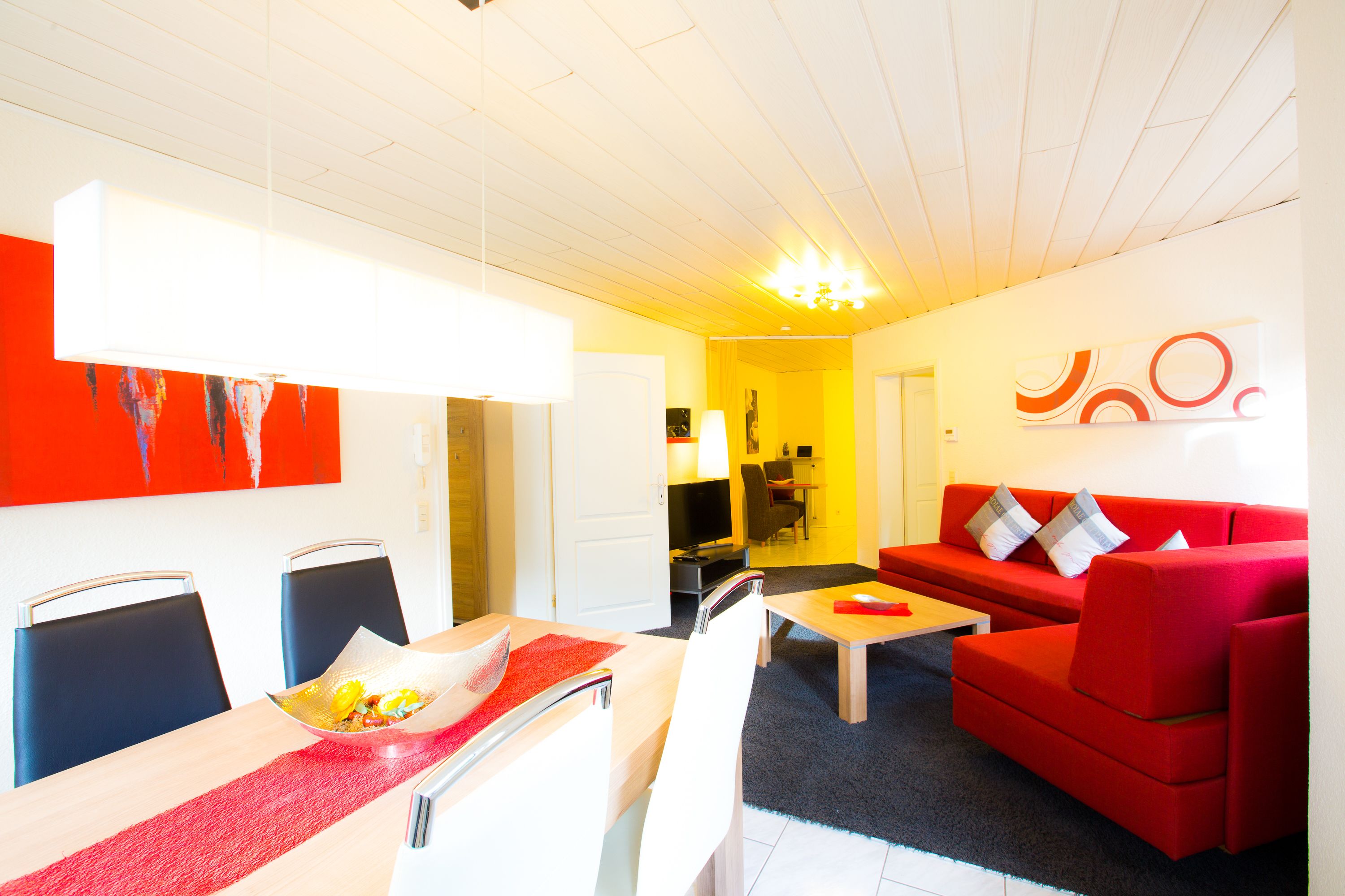 A modern living room with a red sofa, a dining table with chairs, and a TV in the background.