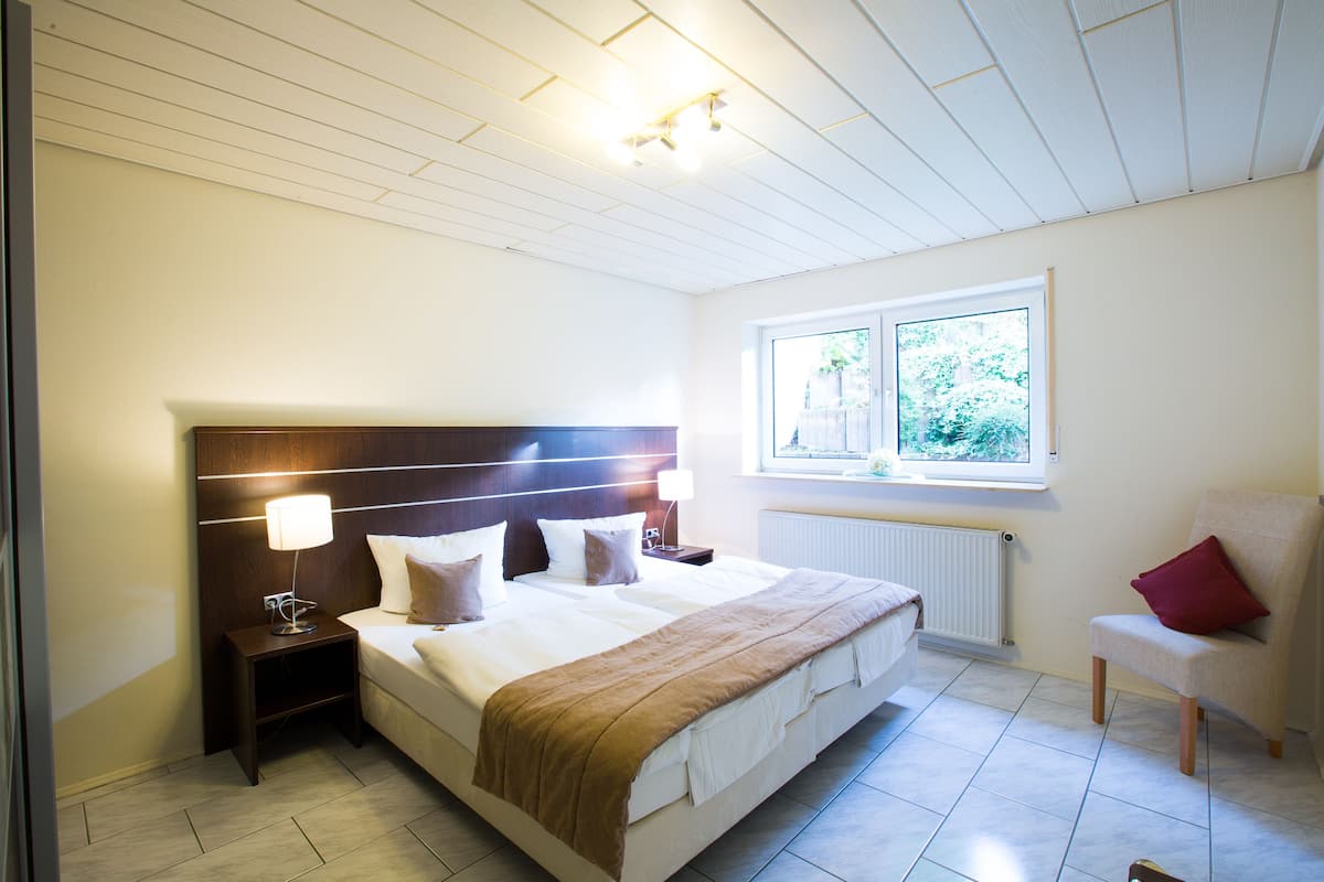 Une chambre avec un grand lit, une table de chevet, une chaise, une fenêtre donnant sur un paysage verdoyant et un plafond doté d’un éclairage encastré.