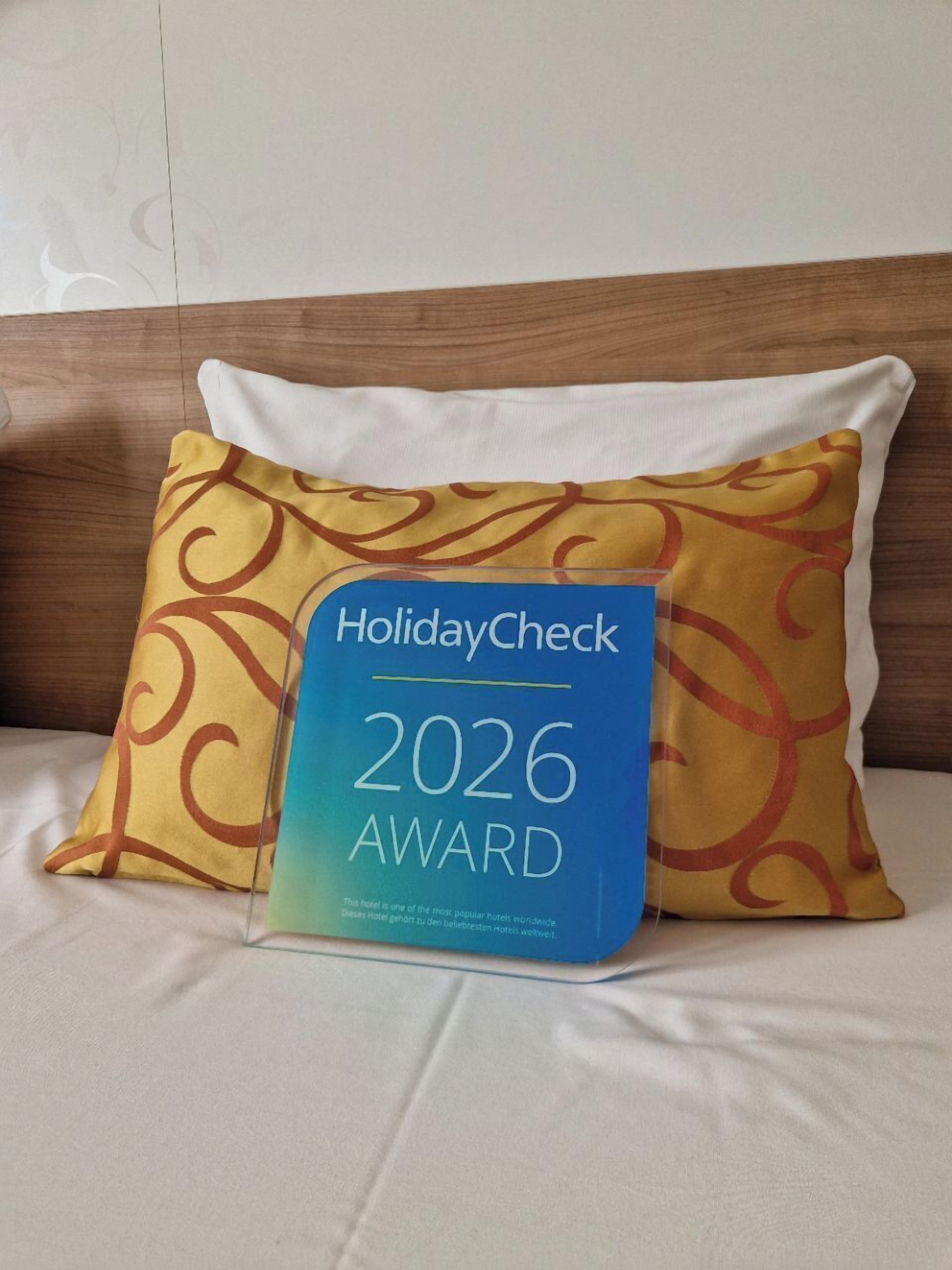 A bed with a white pillow and a pillow with a yellow pattern. In front of the pillow is a blue award plaque with 'HolidayCheck 2026 Award' written on it.