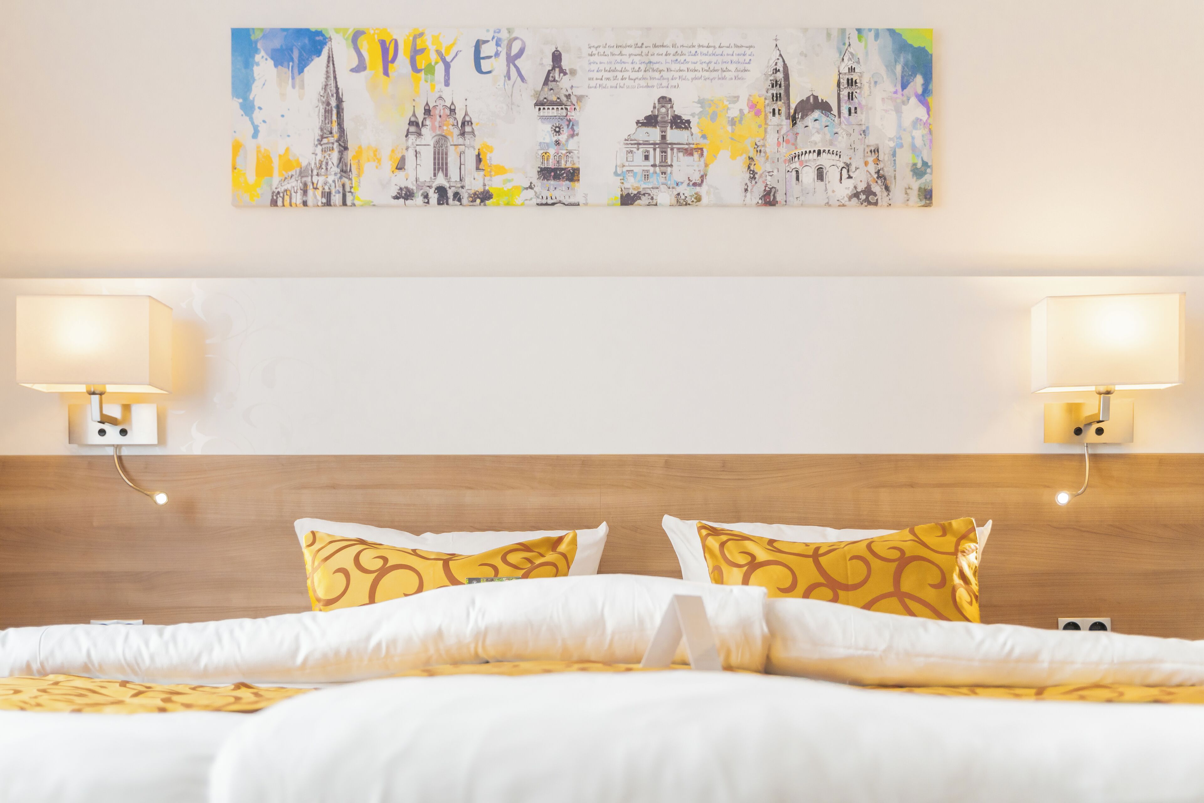 A hotel room with a bed, two wall-mounted lamps, and a cityscape painting on the wall.