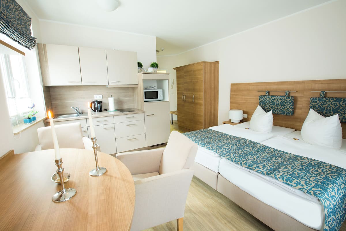 Une chambre d’hôtel moderne avec un lit en bois, une petite kitchenette, un coin repas avec une table et des chaises, et une grande fenêtre.
