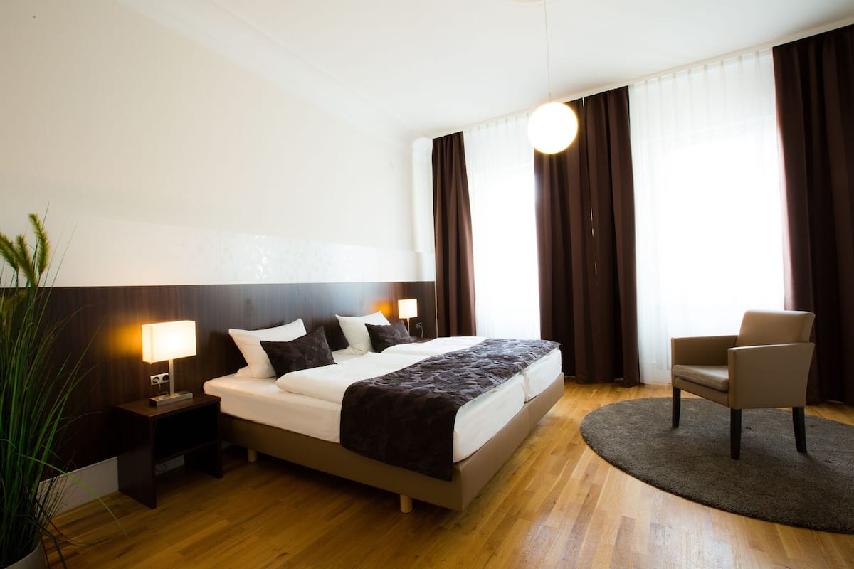 Une chambre d’hôtel moderne avec un grand lit, une table de chevet avec une lampe, une chaise et une grande fenêtre avec des rideaux.