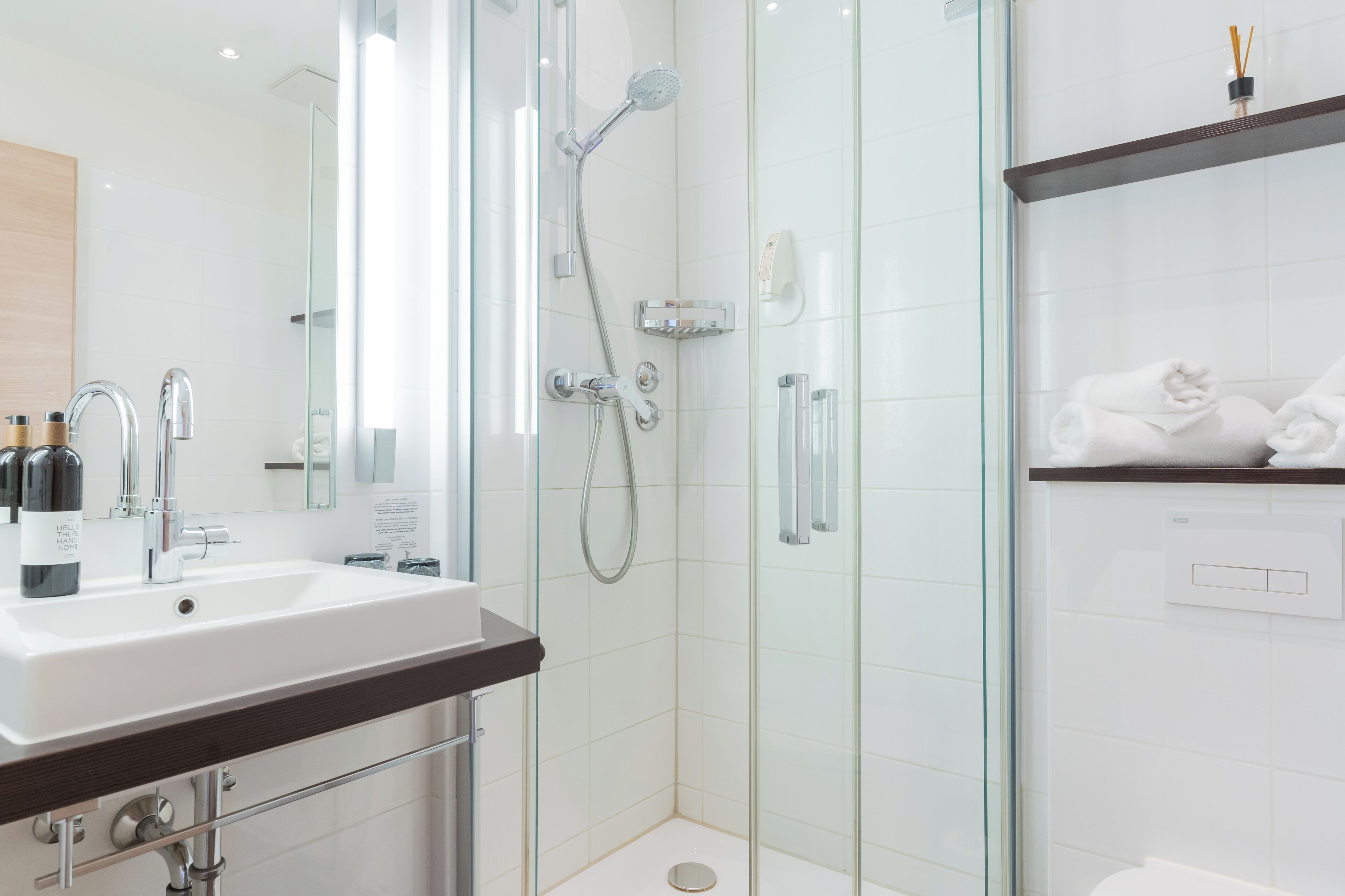 A modern bathroom with a glass-enclosed shower, a white sink with a dark countertop, and a shelf with towels.