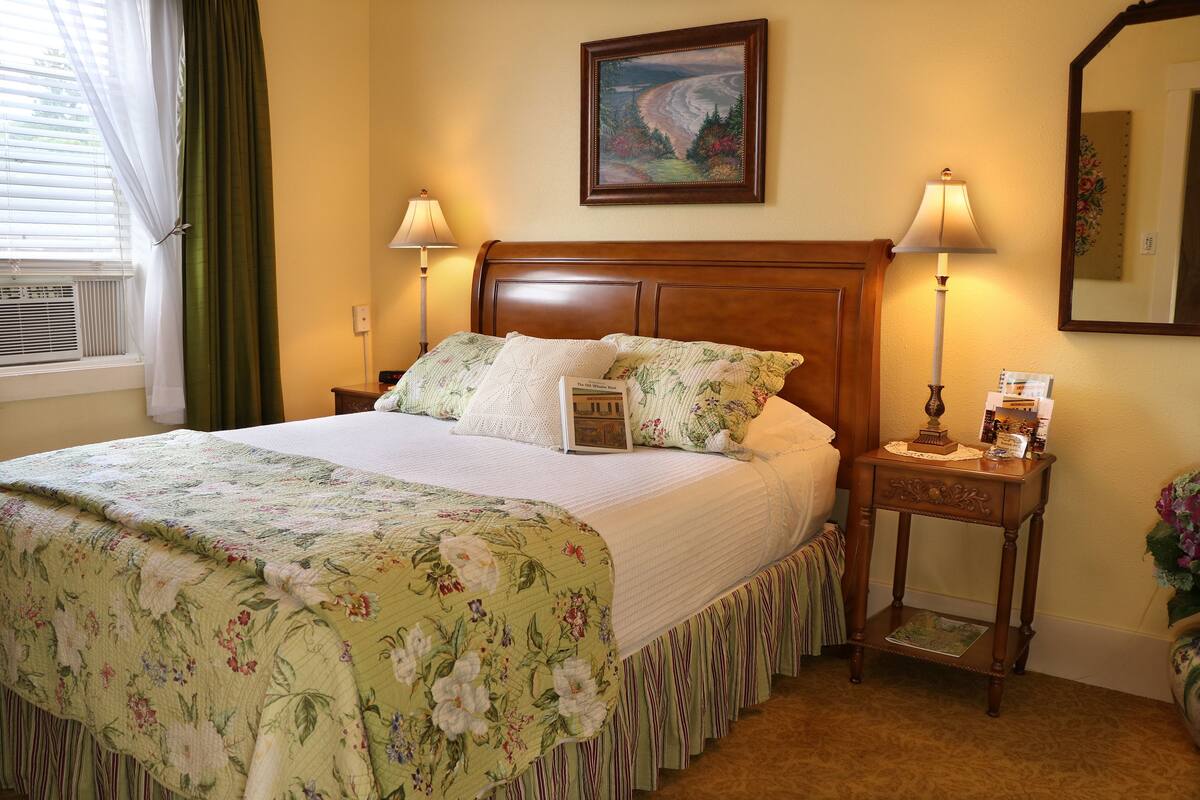Una cama bien hecha con ropa de cama floral, dos lámparas de noche, una mesita de noche con lámpara, un cuadro en la pared y una ventana con cortinas.