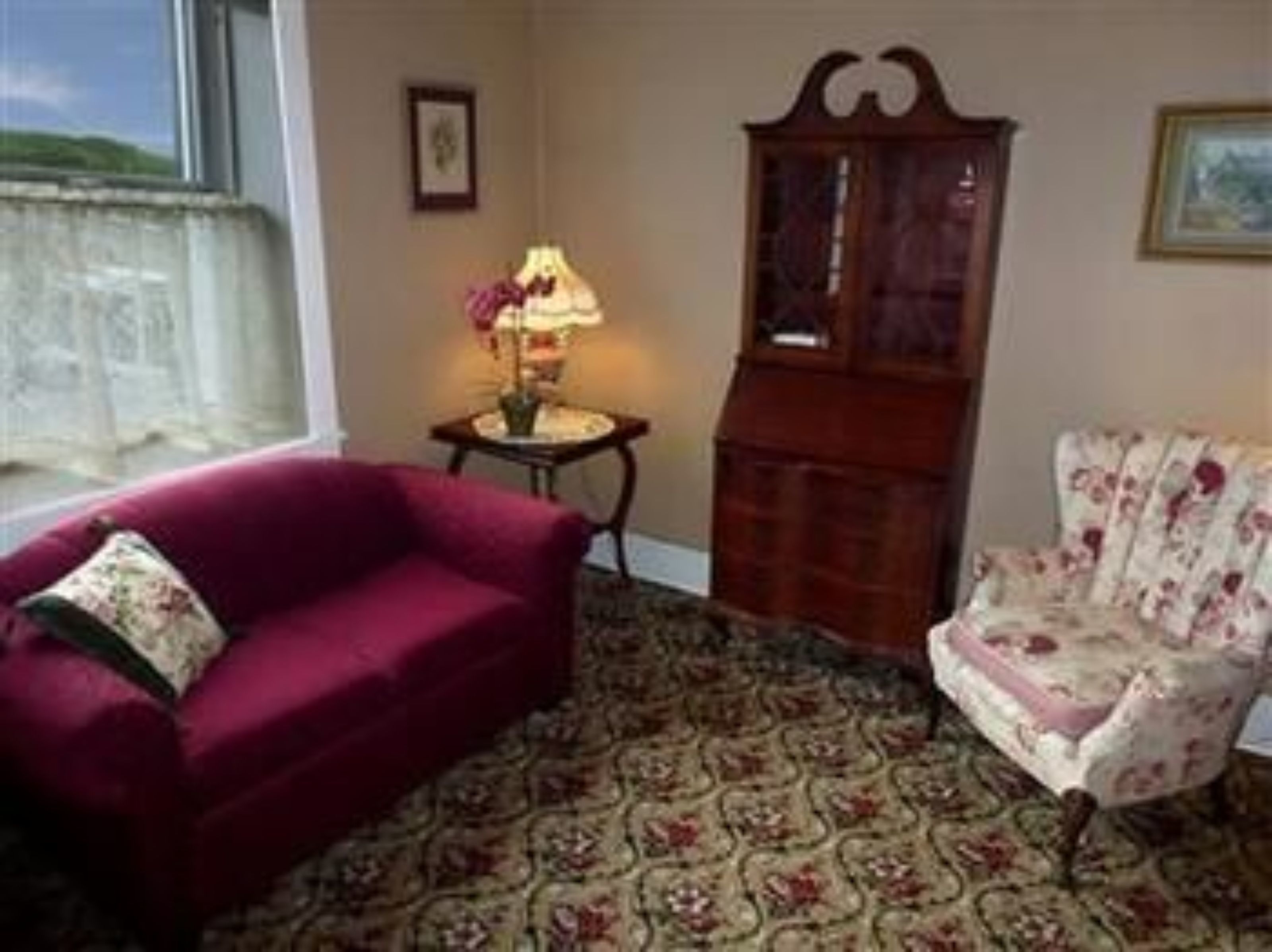 A room with a red sofa, a floral armchair, a wooden cabinet, and a patterned carpet.