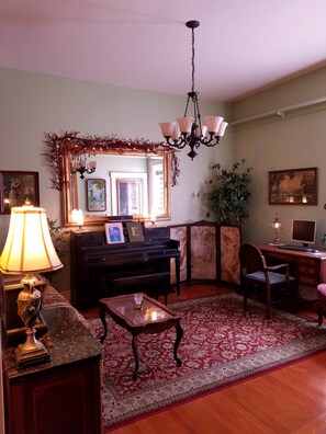 A living room with a piano, a desk with a computer, a chair, a coffee table, a lamp, a mirror, and a chandelier.