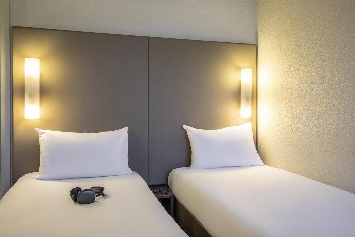 Habitación de hotel con dos camas individuales, cada una con ropa de cama y almohadas blancas, y un par de auriculares sobre la cama.