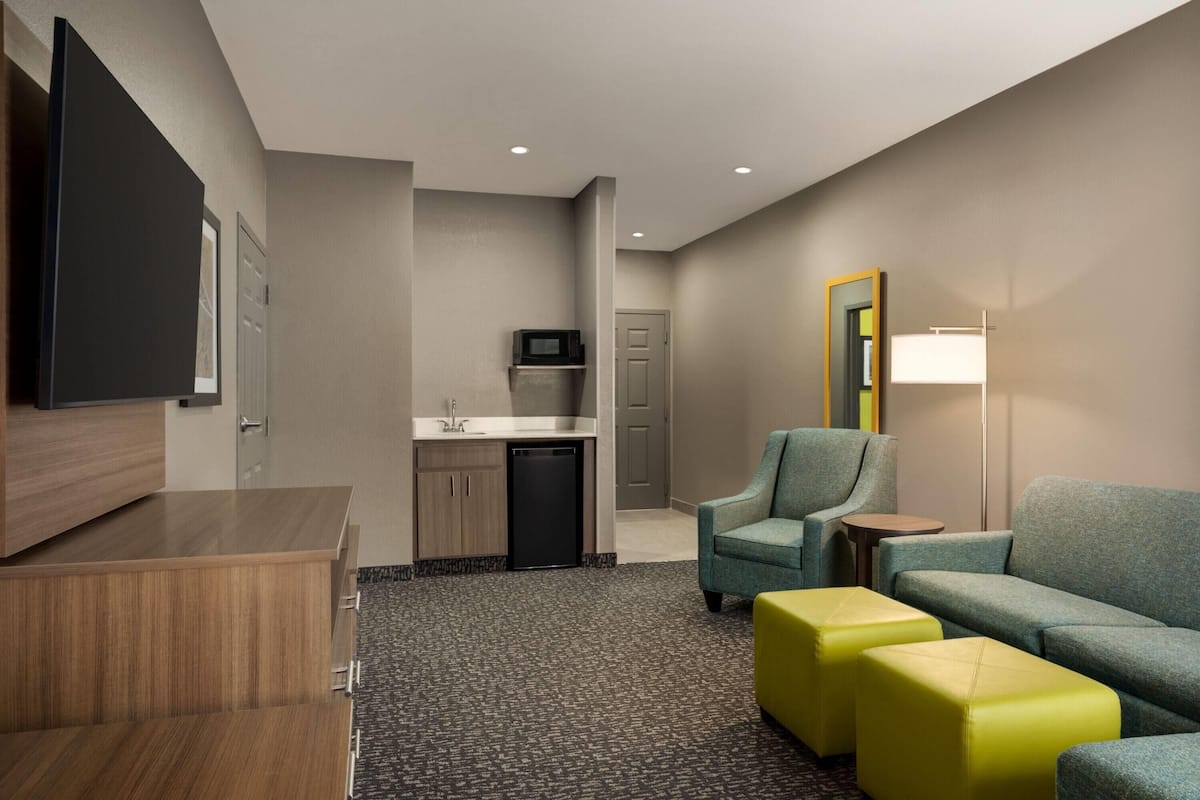 A hotel room with a kitchenette, a seating area with a sofa and ottomans, a television, and a mirror.