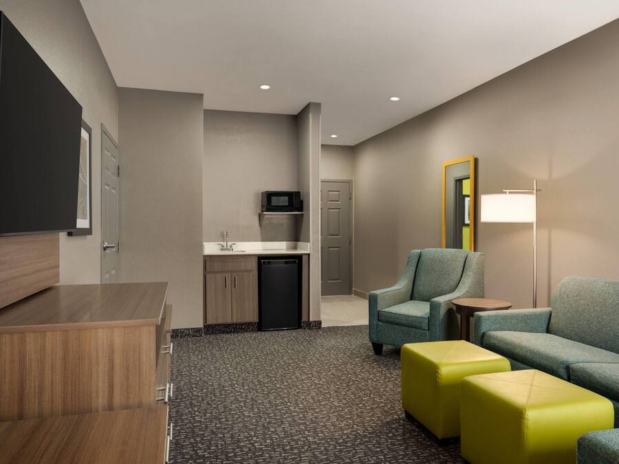 A hotel room with a kitchenette, a seating area with a sofa and ottomans, a television, and a mirror.