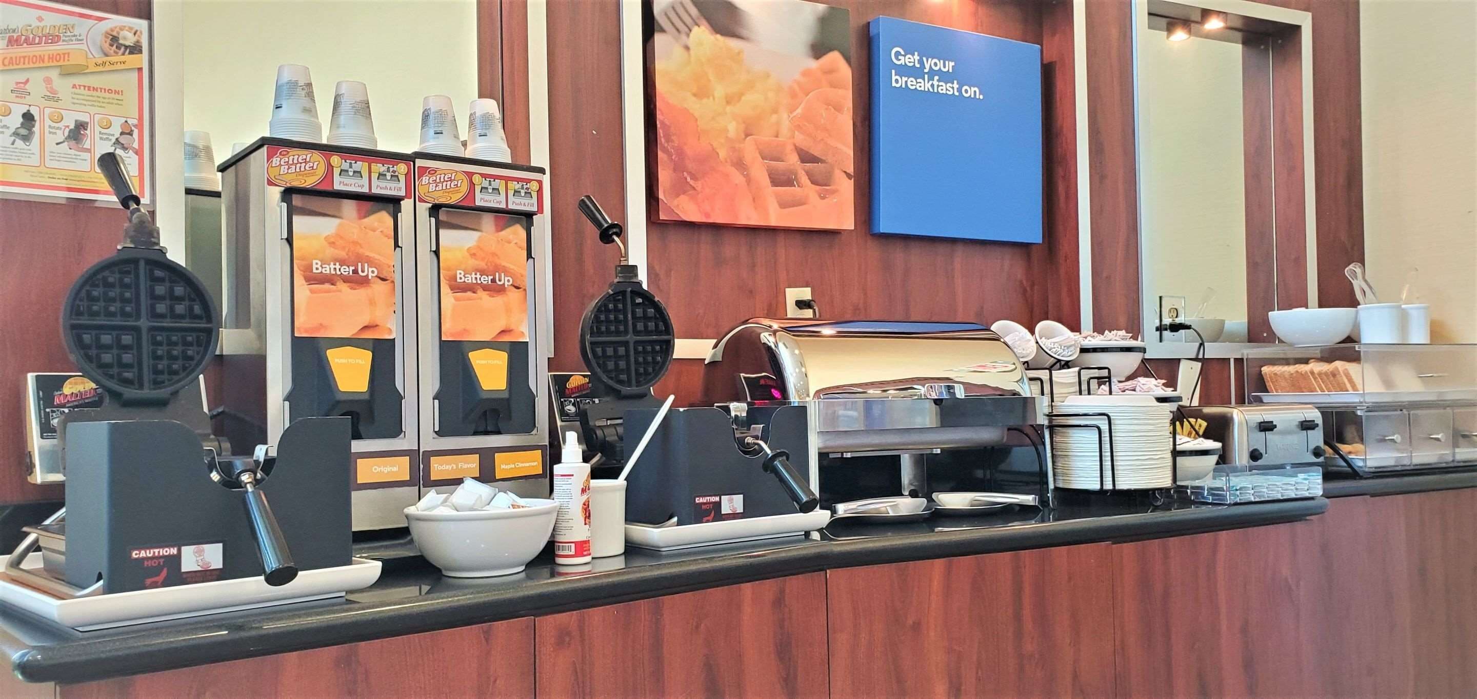 Estación de desayuno con máquinas para hacer gofres, cafeteras y un cartel que anuncia el desayuno.