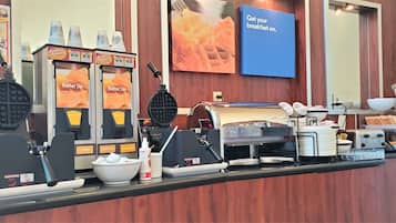 Estación de desayuno con máquinas para hacer gofres, cafeteras y un cartel que anuncia el desayuno.