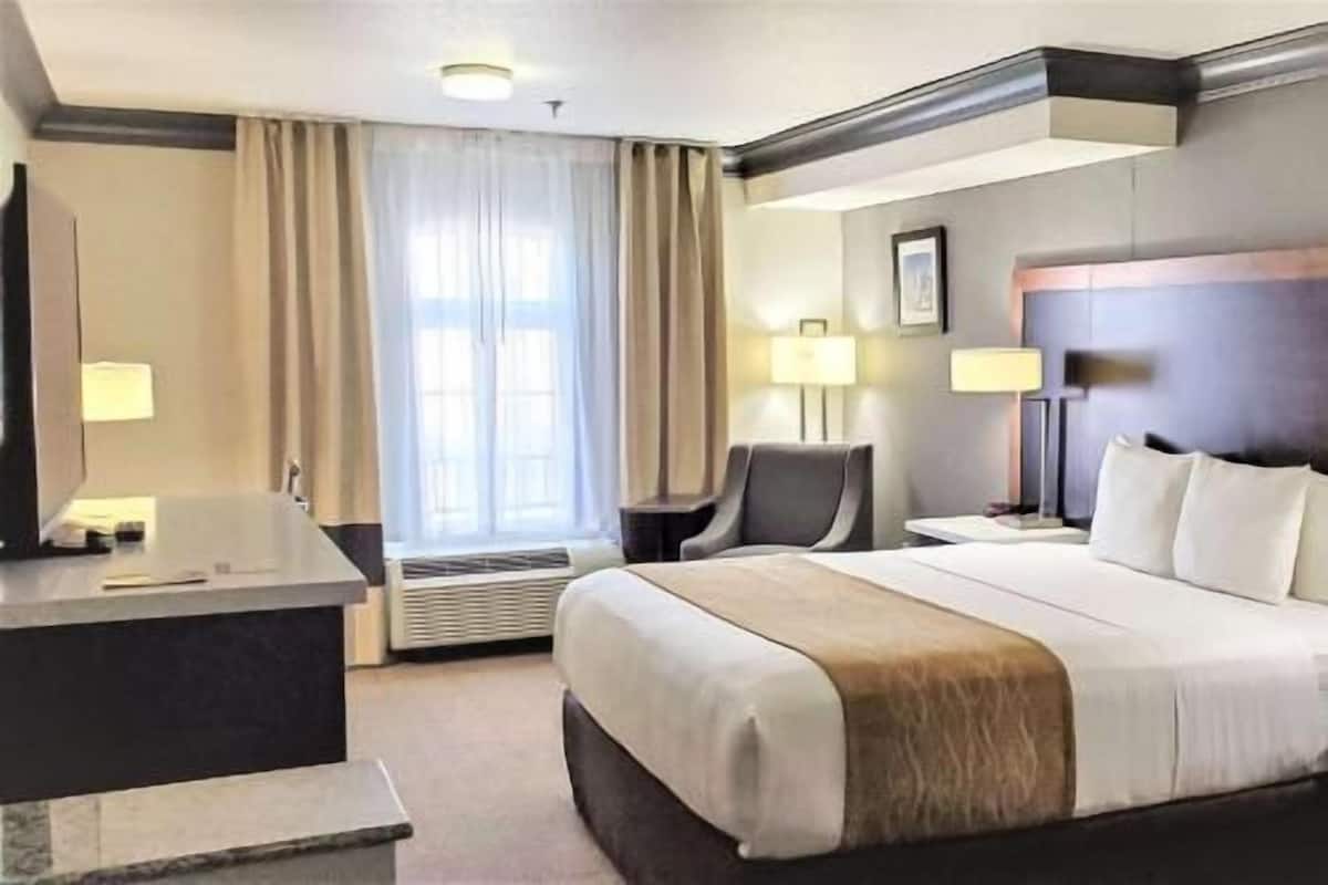 Une chambre d’hôtel comprenant un lit, un bureau, une chaise, deux tables de chevet, une lampe et une fenêtre avec des rideaux.