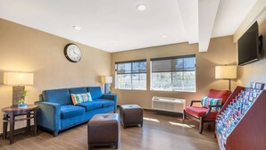 A living room with a blue sofa, brown ottoman, and a red chair. There is a clock on the wall, a flat-screen TV, and a window with blinds.