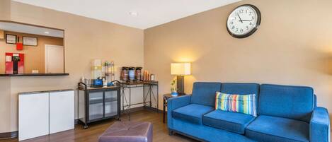 A living room with a blue sofa, a clock on the wall, a small table with a lamp, and a kitchenette area.