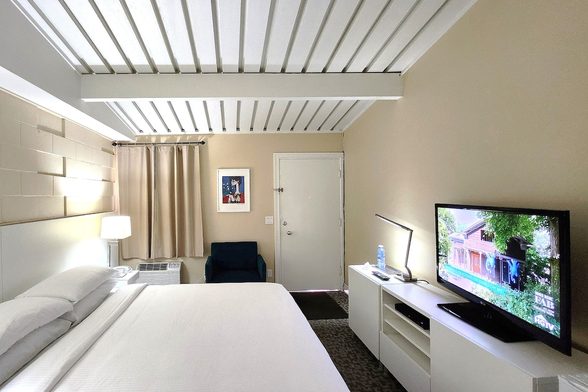 A hotel room with a bed, a desk with a television, a chair, and a window with curtains.