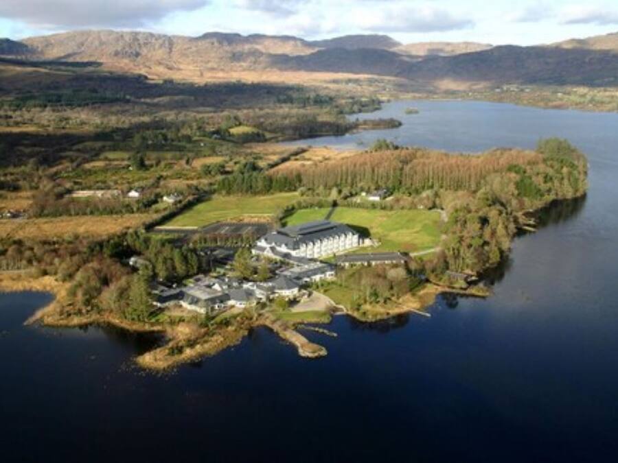 An aerial view of a lakeside property with a large building, surrounded by forested areas and hills in the background.