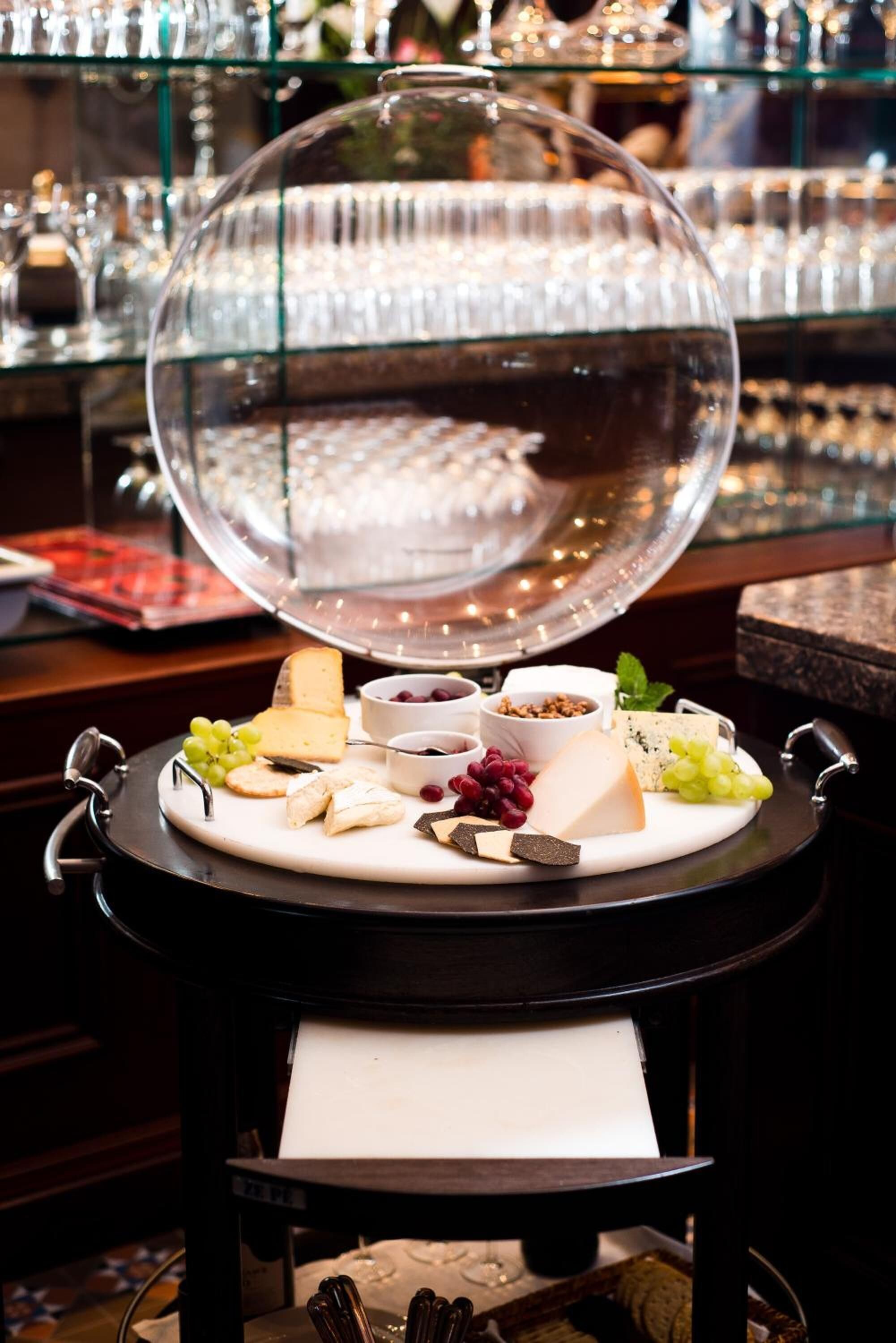A cheese board with various cheeses, fruits, and nuts on a tray, under a glass dome.