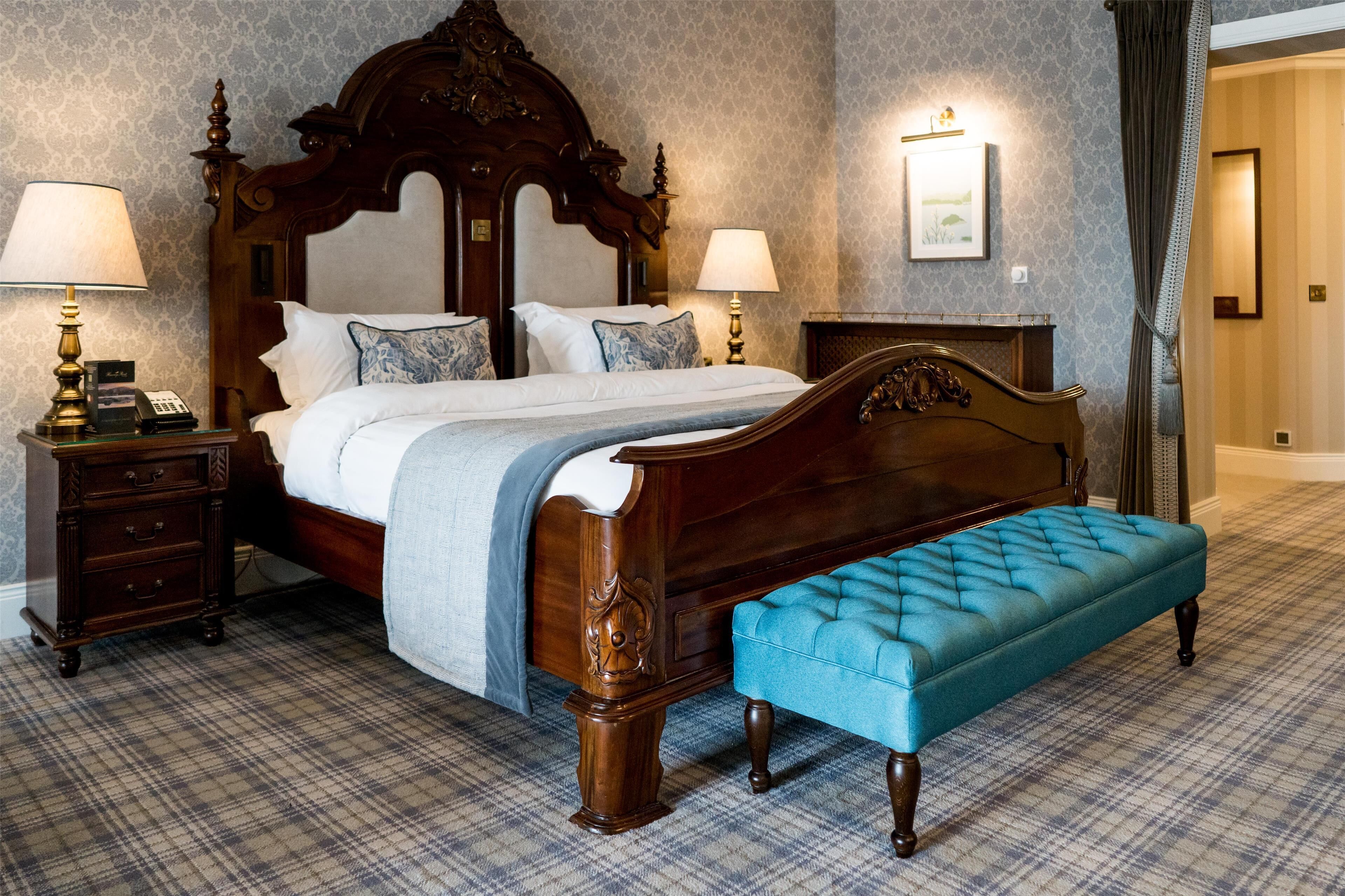 A classic four-poster bed with a tufted bench, two bedside tables with lamps, and a plaid carpet.
