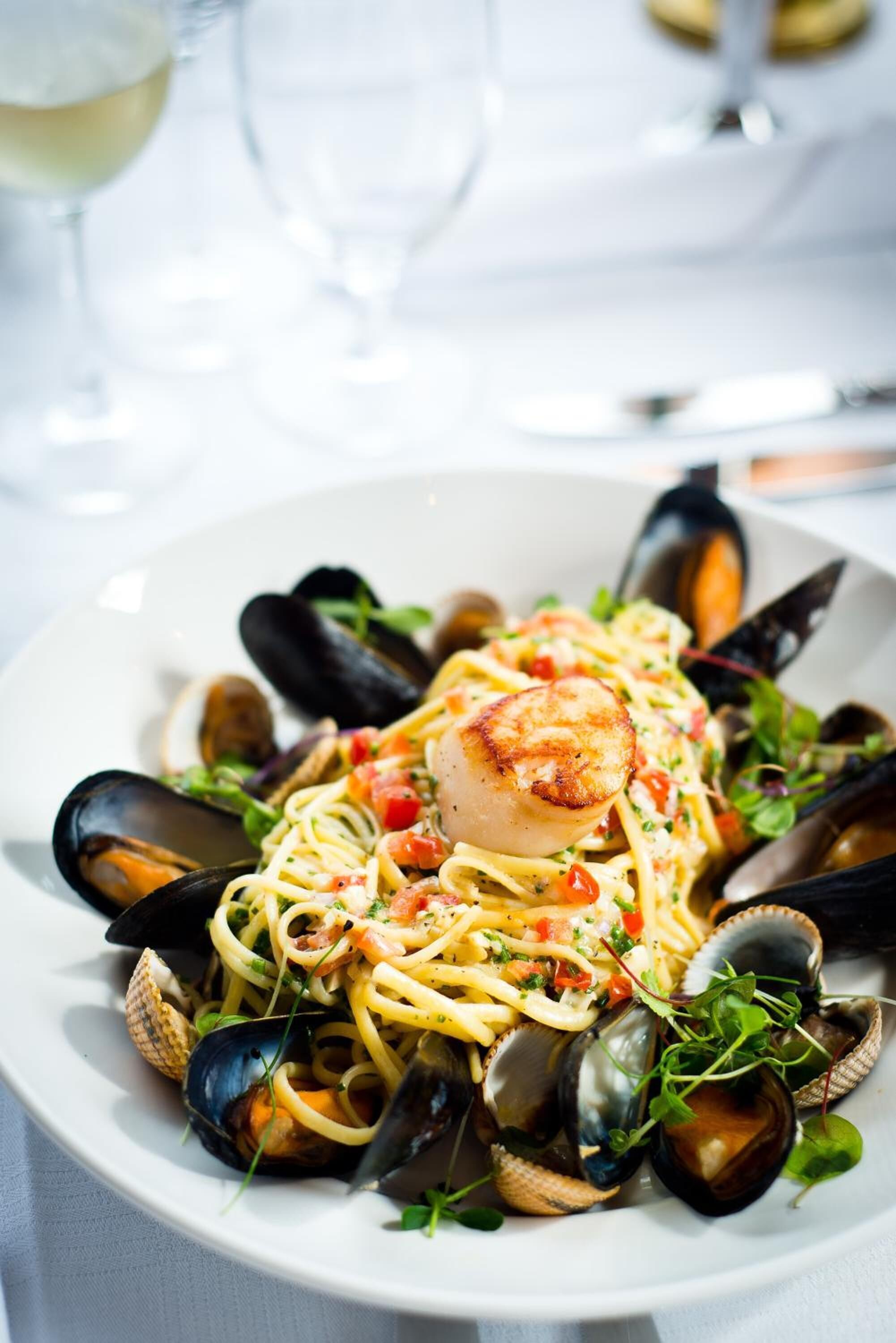 A seafood pasta dish with mussels and a seared scallop on a white plate.