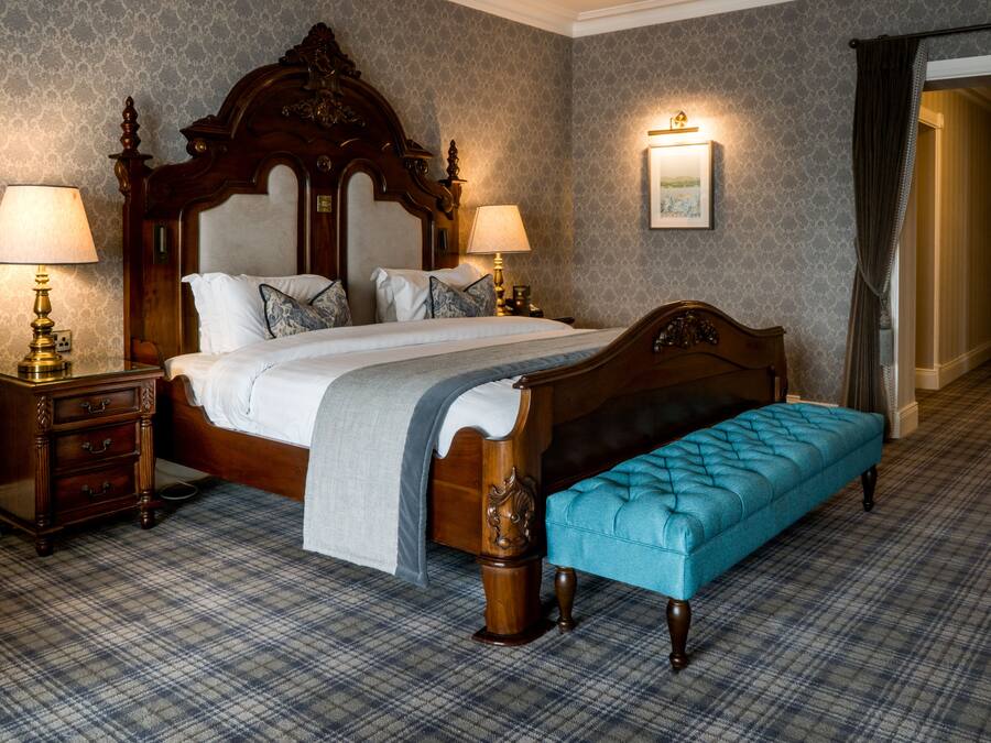 A neatly made bed with a wooden headboard, two bedside tables with lamps, a blue upholstered bench, and a plaid carpet.