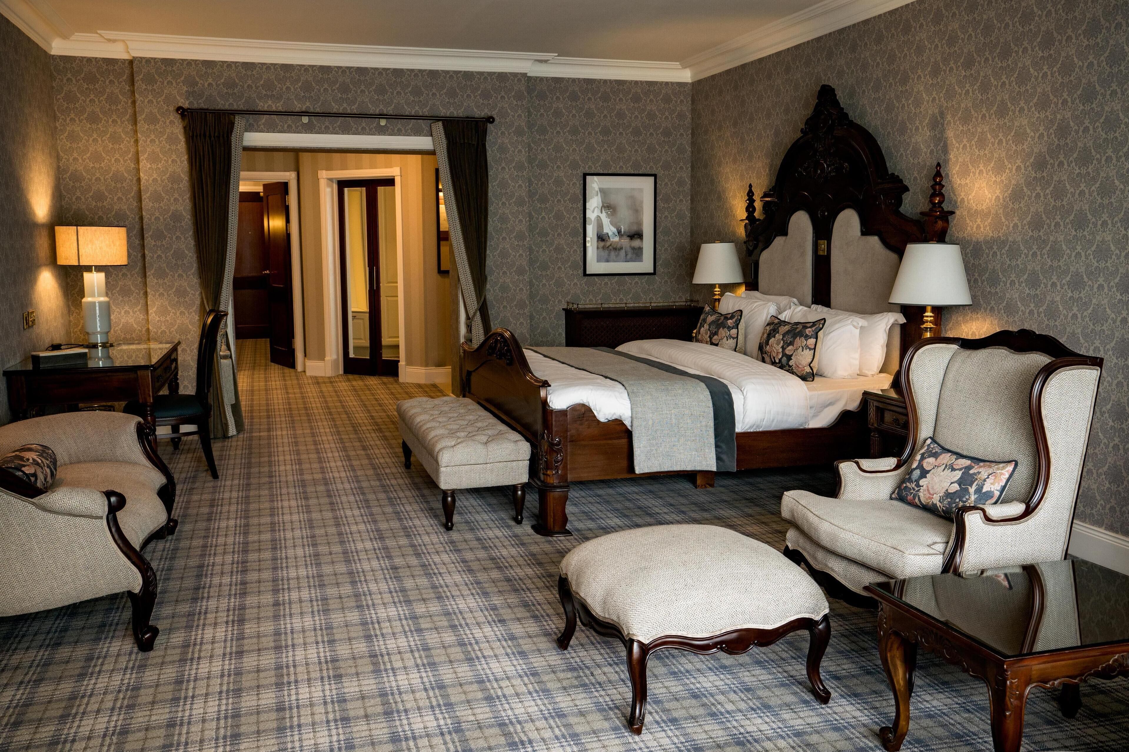 A hotel room with a large bed, two armchairs, a small table, and a bench.