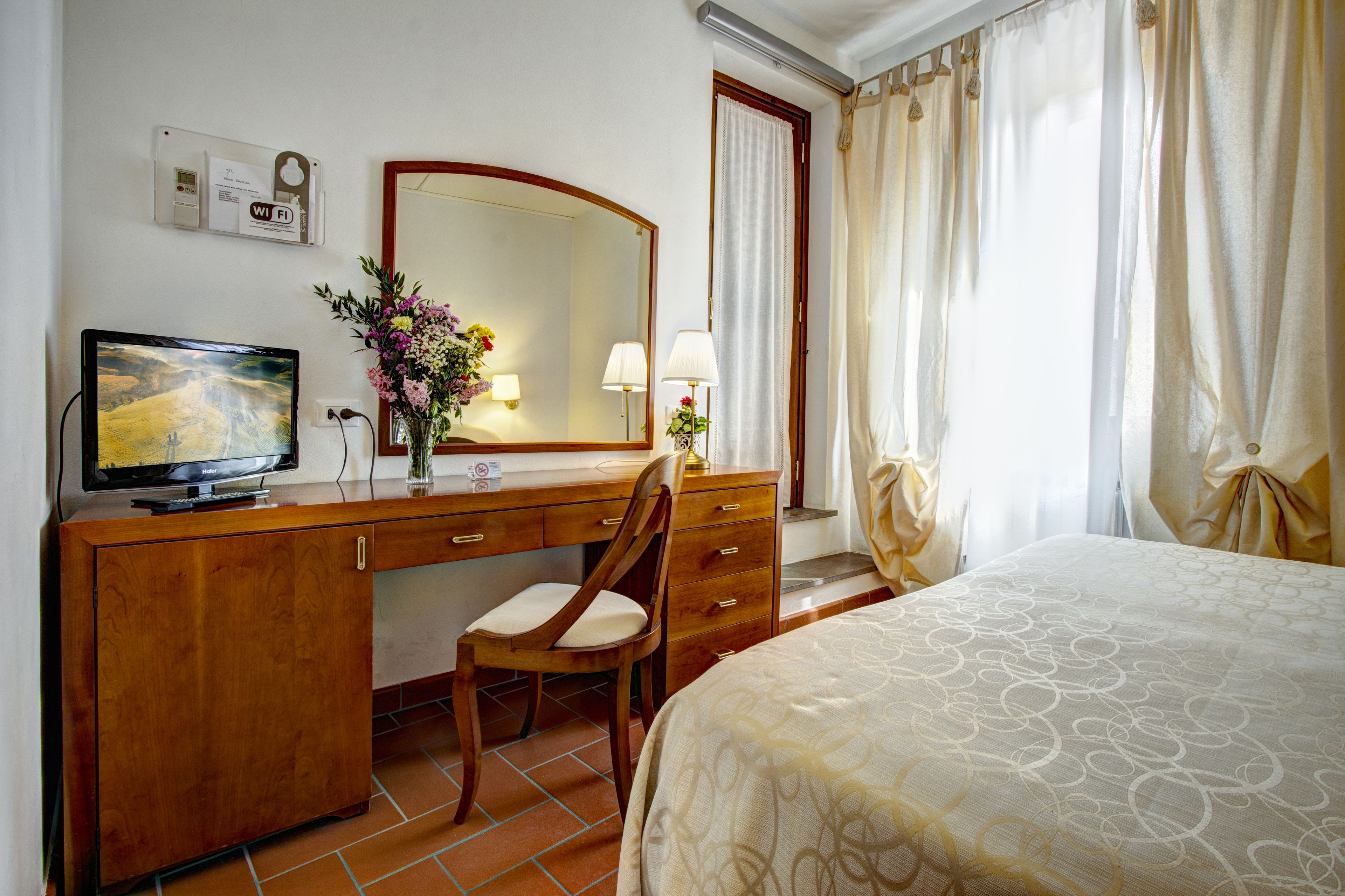 Habitación de hotel con escritorio de madera, una silla, cama, televisor, espejo y ventana con cortinas.