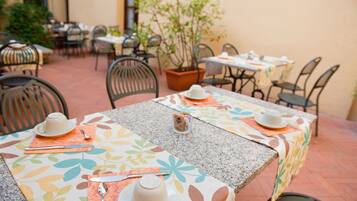 An outdoor dining area with tables set for two, featuring floral tablecloths and a potted plant in the center.
