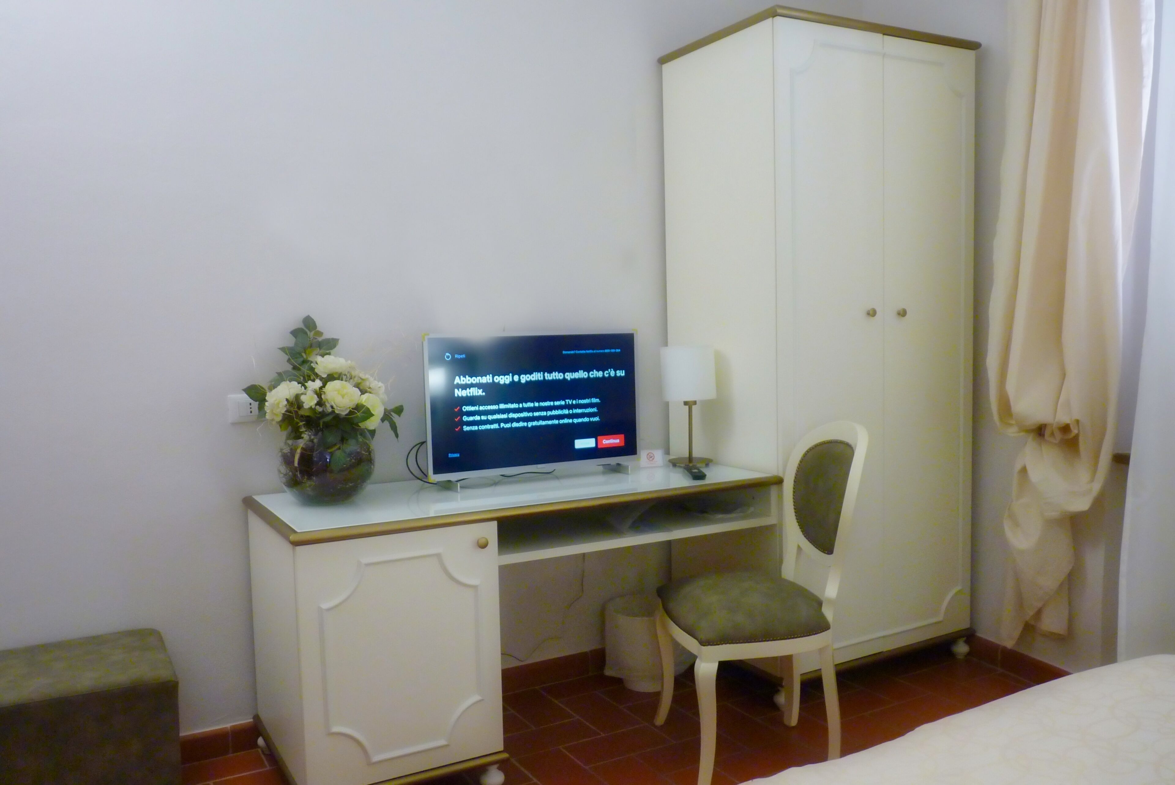 A hotel room with a desk, a chair, a television, a vase with flowers, and a wardrobe.
