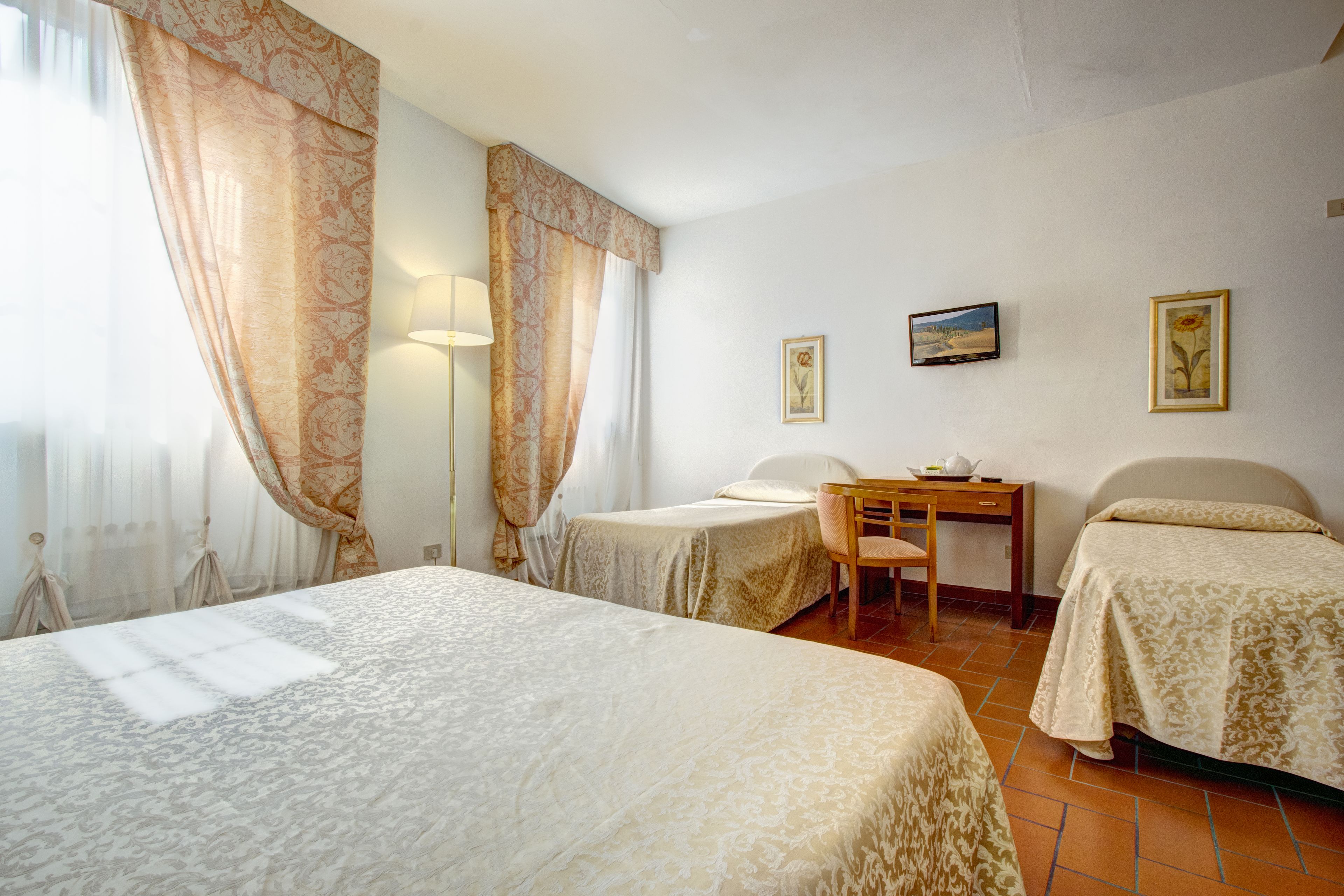 Habitación de hotel con dos camas, un escritorio y una silla. Hay cortinas y dos cuadros en la pared.