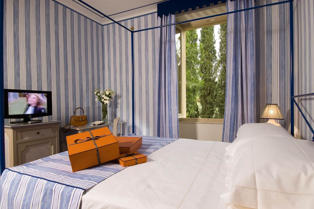 Una habitación de hotel con papel tapiz a rayas, una cama con sábanas blancas, una caja de regalo naranja, un televisor en una cómoda, un florero con flores y una ventana con cortinas.